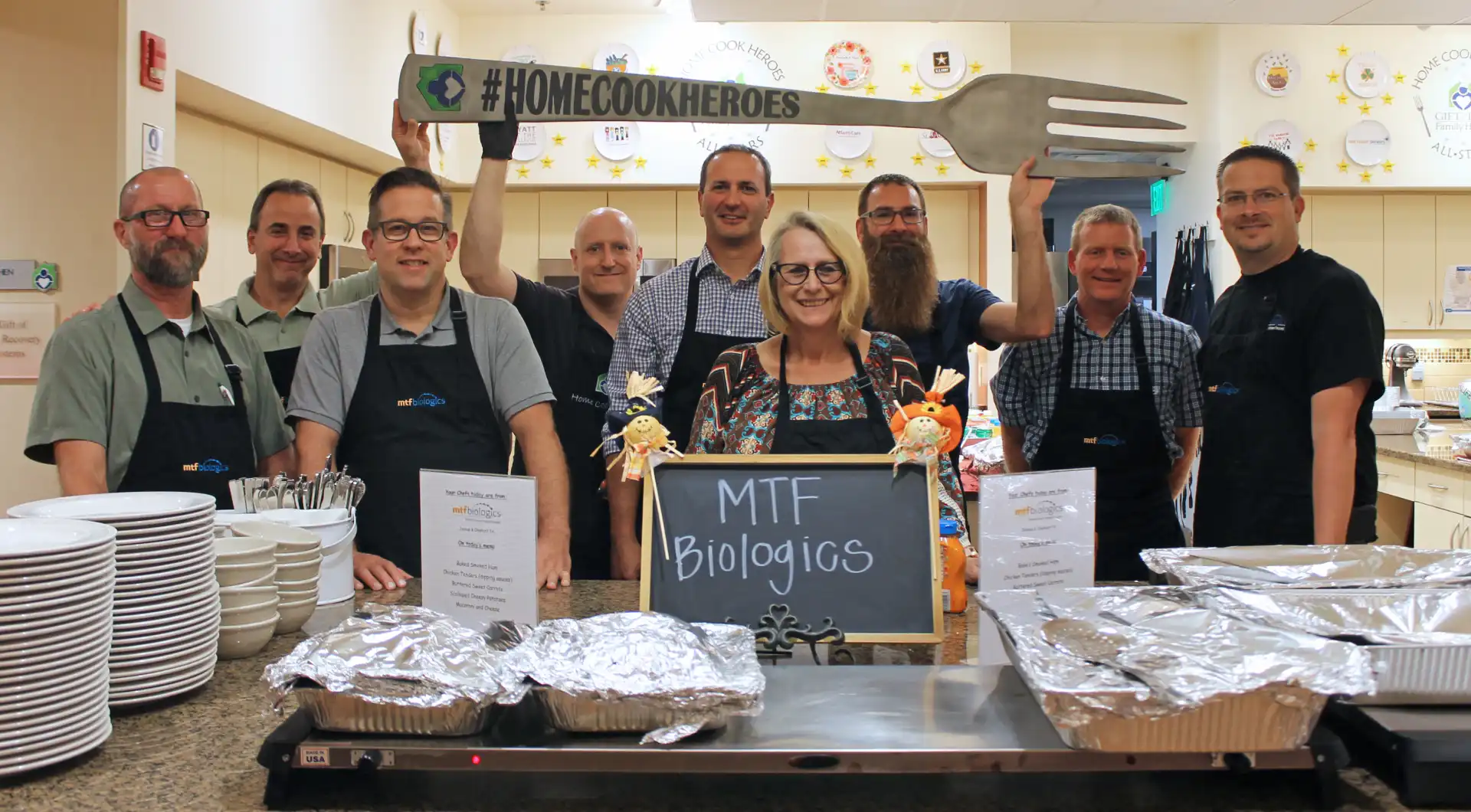 Group of MTF Biologics volunteers smiling in a kitchen, holding a large fork with a sign reading #HomeCookHeroes while serving food at Howie's House